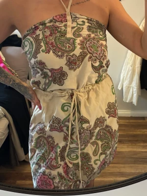 Mönstrad axelbandslös sommarklänning - Snygg axelbandslös kortklänning med paisleymönster i grönt, rosa och beige. Klänningen har knappar framtill, snörning i midjan och är perfekt för varma dagar. Tillverkad i ett mjukt bomullstyg som känns skönt mot huden. Passar perfekt till en dag på stranden 