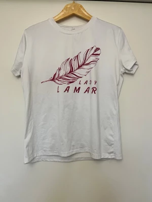 Vit Lady Lamar t-shirt med fjäder - Vit t-shirt från Lady Lamar med ett tryck av en röd fjäder och texten 'Lady Lamar' på bröstet. Klassisk rund halsringning och korta ärmar. Tillverkad i mjuk bomull för en bekväm känsla.