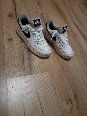 Nike Air Force 1 vita sneakers - Klassiska vita Nike Air Force 1 sneakers med svart swoosh på sidan. Skorna har perforerad tåbox för ventilation, vit sula och vita skosnören. Tillverkade i läder med textildetaljer och låg profil som ger en clean och tidlös look.