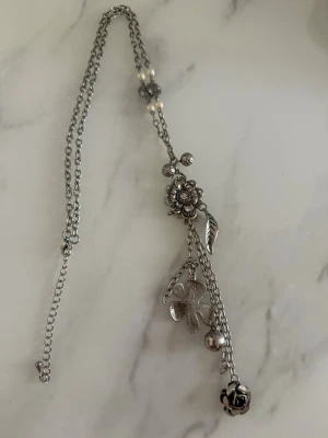 Silverfärgat halsband med blommor och pärlor - Unikt halsband i silverfärg med flera hängen i form av blommor, blad och en liten klocka. Kedjan har små vita pärlor och detaljerade metallhängen som ger en bohemisk och cool vibe. Perfekt för dig som gillar smycken med mycket detaljer och naturinspirerade former.