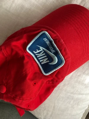 Röd keps från Nike Sportswear - Snygg röd keps från Nike Sportswear med ett blått och vitt broderat märke framtill. Klassisk böjd skärm och justerbar rem bak. Tillverkad i bomull för en bekväm och avslappnad look. Perfekt accessoar för en sportig stil.