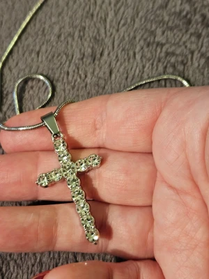 Silverfärgat halsband med korshänge - Snyggt halsband i silverfärg med ett korsformat hänge dekorerat med gnistrande klara stenar. Kedjan är tunn och smidig, och korset har en modern, stilren design som verkligen drar blickarna till sig. Perfekt accessoar för dig som gillar bling och vill sticka ut.