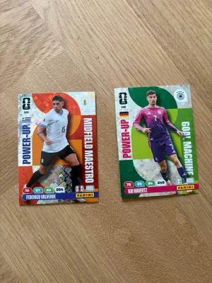 Panini fotbollssamlarbilder - Två Panini fotbollssamlarbilder med holografisk effekt. Ena kortet har röd och orange bakgrund med texten 'Midfield Maestro', det andra har grön och silver bakgrund med texten 'Goal Machine'. Båda visar fotbollsspelare i matchställ och har statistik och namn tryckt på framsidan.