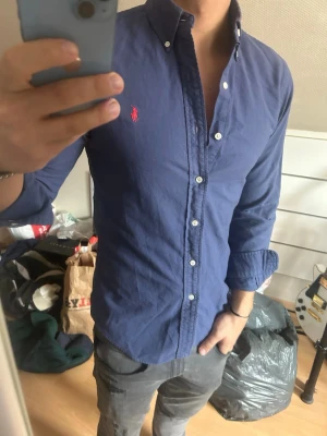 Blå skjorta från Ralph Lauren - Säljer en klassisk blå skjorta från Ralph Lauren med knappkrage och vita knappar. Skjortan har lång ärm och den ikoniska röda loggan broderad på bröstet. Perfekt för dig som gillar stilrena plagg med snygg passform.