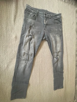 Grå dsquared2 - Säljer ett par grå jeans med slitningar och stora hål på båda benen. Jeansen har klassisk femficksmodell och dragkedja. Perfekta för dig som gillar en edgy och avslappnad stil.