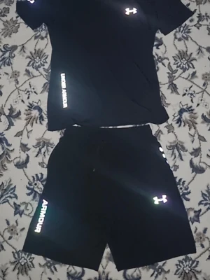 Svart träningsset från Under Armour - Säljer ett svart träningsset från Under Armour med t-shirt och shorts. Setet har vita logotyper och tryck på både tröjan och shortsen. Tillverkat i ett lätt och snabbtorkande material som passar perfekt för gym eller löpning.