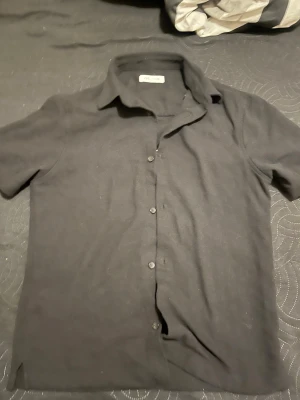 Svart kortärmad skjorta med knappar - Stilren svart skjorta från Zara med korta ärmar och klassisk krage. Skjortan har knäppning framtill och är tillverkad i ett mjukt bomullsmaterial som känns bekvämt mot huden. Perfekt för en clean och enkel look.