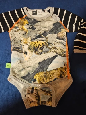 Body med lodjursmotiv från Molo - Cool body från Molo med svartvita ränder på ärmarna och färgglatt lodjursmotiv på framsidan. Orange sömmar längs sidorna och tryckknappar vid axel och gren. Mjuk bomullskänsla och unik design som sticker ut.