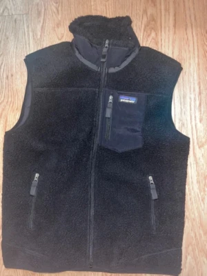 Svart fleeceväst från Patagonia - Mysig svart fleeceväst från Patagonia med hög krage och hel dragkedja. Västen har tre fickor med dragkedja, varav en bröstficka med logga. Tillverkad i mjukt teddyfleece-material som håller värmen och ger en avslappnad look.