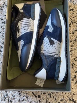 Valentino Garavani blå sneakers - Snygga sneakers från Valentino Garavani i marinblått och vitt med coolt kamouflagemönster. Ovandelen är i mesh och mocka, med detaljer i läder och nitar på sulan. Perfekta för dig som vill sticka ut med stilrena och exklusiva skor.                           SKRIV INNAN DU KÖPER