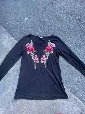 Svart topp med broderade blommor - Svart långärmad topp med v-ringning och broderade blommor i rosa och rött framtill. Toppen har en dekorativ detalj vid halsen och är tillverkad i ett mjukt, tunt material som känns skönt mot huden. Perfekt för dig som gillar unika detaljer och blommiga inslag.