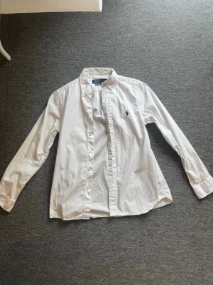 Vit skjorta från Polo Ralph Lauren - Klassisk vit skjorta från Polo Ralph Lauren med lång ärm och knappar framtill. Skjortan har den ikoniska broderade loggan på bröstet och är tillverkad i mjuk bomull. Perfekt till många olika stilar och enkel att matcha. Det är 16 år men det blir som en xs.