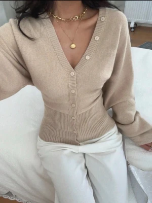 Beige ribbad kofta med v-ringning - Trendig beige kofta med djup v-ringning och ribbad midja som framhäver figuren. Knäppning framtill med matchande knappar och långa ärmar. Perfekt att styla med smycken och vida byxor för en clean look.