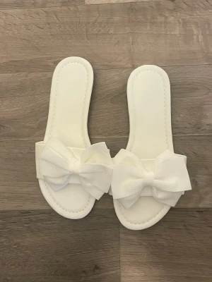 Vita sandaler med rosetter - Snygga vita sandaler med bred rem och stora dekorativa rosetter framtill. Tillverkade i ett slätt material med mjuk innersula för extra komfort. Perfekta för en trendig och feminin look under varma dagar.