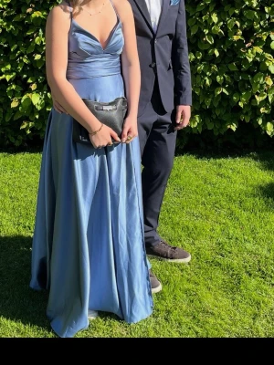 Lång blå satinklänning med smala axelband - Elegant långklänning i glansigt blått satinmaterial med smala axelband och v-ringad omlottringning. Klänningen har en figurnära överdel och ett utsvängt fall som ger en lyxig känsla. Perfekt för bal eller student. Om man är intresserad så finns också möjligheten att köpa till näsduken som är i samma blå färg, som ni ser på bilderna. Den har korsett på baksidan som såklart gör klänningen justerbar!