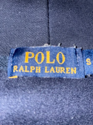 Marinblå hoodie från Polo Ralph Lauren - Snygg marinblå hoodie från Polo Ralph Lauren med dragkedja framtill och vit logga på bröstet. Hoodien har vita dragsnören i huvan och två fickor framtill. Perfekt för en avslappnad och stilren look.