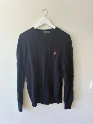 Mörkblå kabelstickad tröja Polo Ralph Lauren - Kabelstickad mörkblå tröja från Polo Ralph Lauren. Tröjan har rund halsringning, långa ärmar och ribbade muddar. Perfekt för alla tillfällen även lite finare. Tröjan är i storlek medium och är väldigt skön och lättburen.