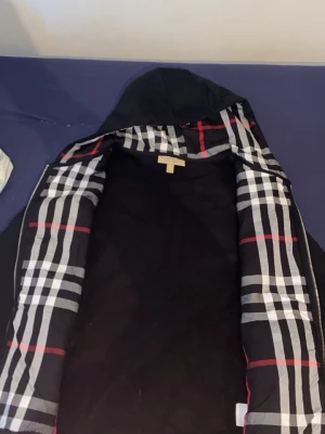 Svart hoodie med rutigt foder Burberry - Säljer en svart hoodie från Burberry med dragkedja och klassiskt rutigt foder i huvan. Hoodien har dragsko i huvan och två fickor framtill. Perfekt för dig som gillar stilrena plagg med en twist av mönster. Materialet känns mjukt och bekvämt.