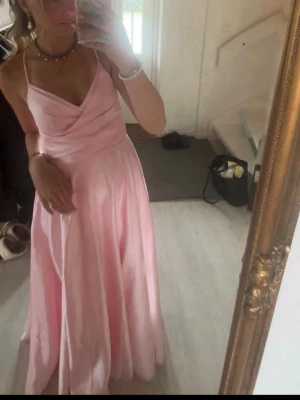 Balklänning - Elegant ljusrosa långklänning/ balklänning med tunna axelband och draperad överdel. Klänningen har en v-ringad urringning och ett flytande, utsvängt fall. Perfekt för bal eller student. Materialet ser ut att vara satin eller polyester för en glansig finish., använd en gång och som ny! Nypris 2000