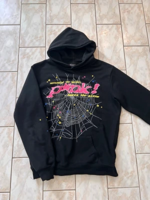 Svart hoodie från Sp5der  - Svart hoodie från Sp5der med färgglatt spindelnäts- och stjärntryck samt texten 'p*nk!' i rosa på bröstet. Tillverkad i 100% bomull och har en klassisk huva och magficka. Perfekt för dig som gillar streetwear och unika prints.