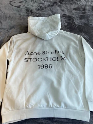 Acne Studios Hoodie Dust White - Säljer en stilren dust vit hoodie med huva och långa ärmar. Hoodien har tryck på baksidan eller logga och har en relaxed fit. Materialet känns mjukt och skönt, perfekt för chill dagar. Passar dig som gillar minimalistisk stil
