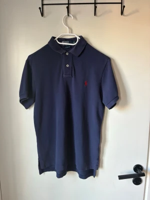 Marinblå pikétröja från Ralph Lauren - Klassisk marinblå pikétröja från Ralph Lauren med korta ärmar och två knappar vid kragen. Tröjan har den ikoniska röda logotypen broderad på bröstet och är tillverkad i bomull för en bekväm känsla. Priset diskutabelt vid snabb affär. Kontakta vid frågor!