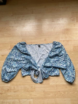 Blommig blå omlottopp från SHEIN - Söt blommig omlottopp från SHEIN i blå toner med knyt framtill. Toppen har långa puffärmar och croppad passform, perfekt för en fräsch och trendig look. Lätt och luftig, passar till både jeans och kjol.