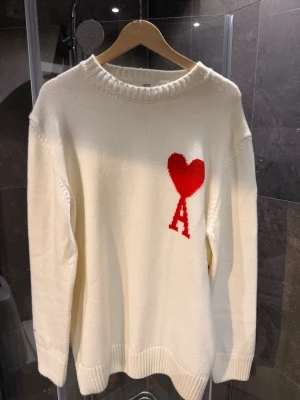 Vit stickad tröja Ami Paris hjärta - Vit stickad tröja från Ami Paris med röd hjärtdetalj och bokstaven A på bröstet. Tröjan har rund halsringning, ribbade muddar och är gjord i mjukt material. Tröjan är knappt använd och är i perfekt skick. 
