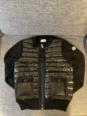 Svart dunväst från Moncler - Snygg svart dunväst från Moncler med glansig, quiltad framsida och två stora fickor med knappstängning. Västen har ribbade muddar och dragkedja framtill. Klassisk Moncler-logga på ärmen och autenticitetsmärkning med taggar kvar.Storleken är 1 vilket motsvarar S