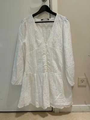  Ny Vit broderad kort klänning med v-ringning - Superfin vit kort klänning med broderade detaljer och v-ringning. Klänningen har långa ärmar och en lätt volang nertill. Perfekt för sommarens alla tillfällen och ger en fräsch och romantisk look.