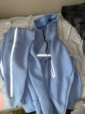 Ljusblå Nike hoodie med dragkedja - Snygg ljusblå tracksuit från Nike med dragkedja framtill och reflekterande detaljer på ärmar och rygg. Hoodien har en stor huva och är tillverkad i ett mjukt, tjockt material som känns riktigt skönt. Perfekt för dig som gillar sportig och modern stil.