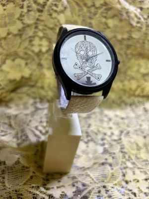 Vit klocka med döskallemotiv - Cool klocka med vit, ormmönstrad rem och svart boett. Urtavlan har ett stort döskalle- och korslagda ben-motiv med detaljer i svart. Perfekt för dig som gillar edgy accessoarer och vill sticka ut lite extra.  Helt ny , Visa i butiken längre, sälja rabatter pris 