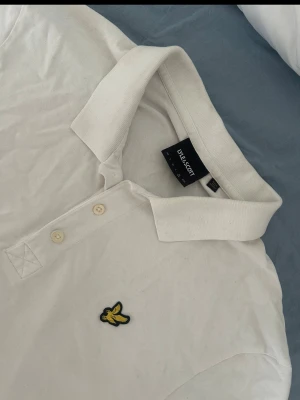 Vit pikétröja från Lyle & Scott - Klassisk vit pikétröja från Lyle & Scott med gul broderad logga på bröstet. Tröjan har krage, korta ärmar och knappar framtill. Tillverkad i mjuk bomull som känns skön mot huden. Perfekt till en clean och enkel stil.
