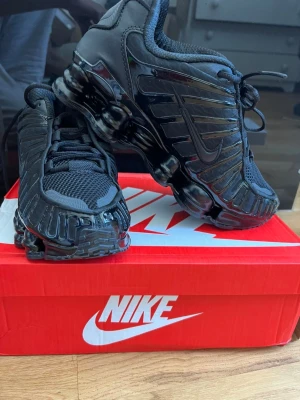 Nike Shox TL helsvarta sneakers - Nike Shox TL sneakers i helsvart med ikoniska fjädrande Shox-sulan. Ovandel i mesh och syntetmaterial med snygga detaljer och ton-i-ton Swoosh-logga. Skorna har en sportig vibe och är perfekta för dig som gillar en futuristisk look.