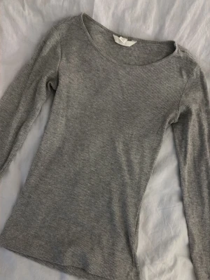 Grå ribbad långärmad topp H&M - En enkel och stilren grå långärmad topp från H&M. Toppen är ribbad och har en rund halsringning. Perfekt att matcha med jeans eller kjol för en clean look.