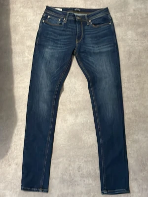 Mörkblå skinny jeans Jack & Jones - Snygga mörkblå skinny fit jeans från Jack & Jones, modell Liam. Jeansen har klassisk femficksdesign, slitningar och kontrastsömmar. Perfekta för dig som gillar en smal passform och tidlös stil.