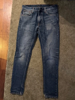 Blå raka jeans med slitningar - Säljer ett par klassiska blåa Nudie Jeans med rak passform och snygga slitningar på framsidan. Modellen är Lean Dean (slim-fit) med färgen troubled sea. Jeansen har fransade benslut och kontrastsömmar. Perfekta för en avslappnad och trendig look. Materialet är denim i bomull. Storleken är W30, L32.
