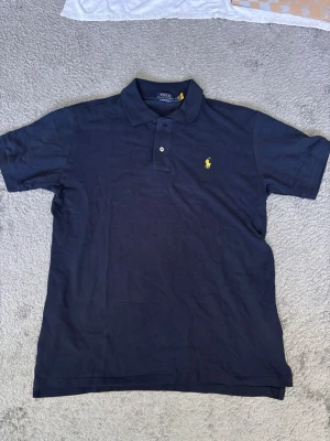 Marinblå pikétröja från Ralph Lauren - Hjälper farsan att sälja av hans garderob. Det är en mörkblå piké med gult Ralph Lauren märke. Han är runt 185 och lite kraftigare. Den passar även en som har L. Skriv om du har funderingar😊