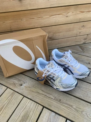Asics vita och silver sneakers - Snygga Asics sneakers i vitt, silver och marinblått med mesh- och syntetmaterial. Skorna har klassisk låg profil, tydliga Asics-loggor på sidorna och en chunky sula för extra komfort. Perfekta för dig som gillar sportig och trendig stil.