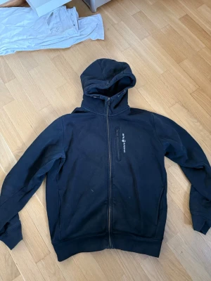 Svart zip-hoodie från sailracing - Svart hoodie med dragkedja från sailracing. Klassisk modell med huva, två sidofickor och logga på bröstet. Tillverkad i mjukt material som känns skönt mot huden. Perfekt för en avslappnad och sportig stil.