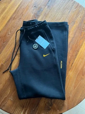 Svarta NOCTA mjukisbyxor från Nike - Snygga svarta mjukisbyxor från Nike NOCTA med gul logga och NOCTA-tryck på benet. Byxorna har dragsko i midjan och är gjorda i mjukt bomullsmaterial för maximal komfort. Perfekta för en chill och avslappnad stil. Storlek S men tvättade en gång så lite mindre 