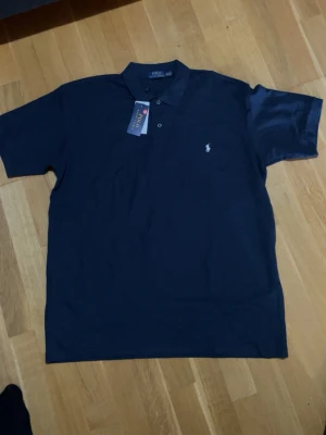 Mörkblå pikétröja från Ralph Lauren - Snygg mörkblå pikétröja från Polo Ralph Lauren med klassisk krage, knäppning framtill och den ikoniska broderade loggan på bröstet. Tillverkad i mjukt bomullsmaterial och perfekt för en stilren look. Komm med bud vid snabb affär 