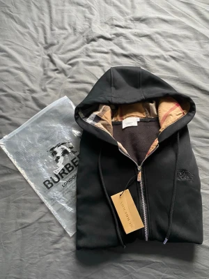 Svart Burberry hoodie med dragkedja - Svart hoodie från Burberry med dragkedja och klassiskt rutigt foder i huvan. Stora fickor framtill, justerbar huva med snören och diskret broderad logga på bröstet. Perfekt för dig som vill ha en stilren och lyxig look.