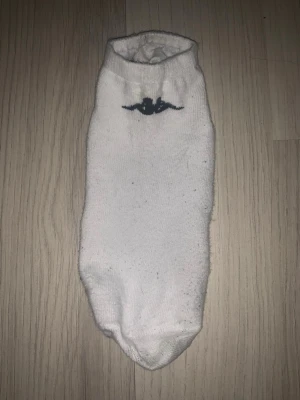 Vita låga strumpor med logga - Ett par vita låga strumpor med en mörkblå logga framtill. Strumporna är enkla och klassiska, perfekta till sneakers eller låga skor. Mjukt material som känns skönt mot huden.