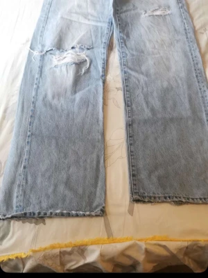 Ljusblåa raka jeans med slitningar - Säljer ett par ljusblå jeans med raka ben och slitna detaljer vid knäna. Jeansen har en avslappnad passform och råa kanter längst ner. Perfekta för en trendig och avslappnad stil.