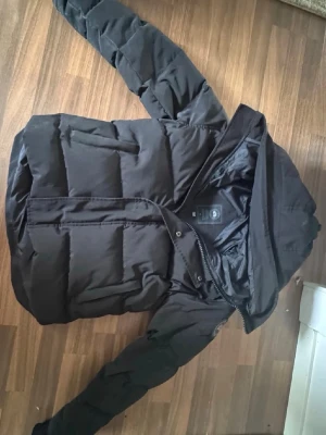 Canada Goose - Puffer Jacket - Säljer en svart pufferjacka med huva och dragkedja framtill. Jackan har breda, vadderade paneler och en klassisk pufferlook. Perfekt för kalla dagar och enkel att matcha med olika outfits.