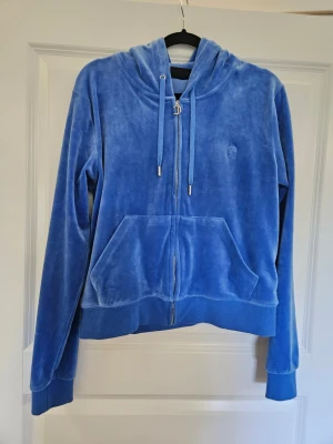 Hoodie Juicy Couture L - Säljer en blå velour hoodie från Juicy Couture med dragkedja framtill, huva med dragsko och fickor på magen. Ikoniskt JC-märke broderat på bröstet och snygga metalldetaljer på snörena. Perfekt för en chill och trendig look.