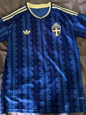 Blå Sverige fotbollströja Adidas - Säljer en blå fotbollströja från Adidas med Sveriges landslagsemblem på bröstet. Tröjan har gula detaljer, klassiska Adidas-ränder på axlarna och ett unikt mönster i olika blå nyanser. Tillverkad i lätt och ventilerande Climalite-material.