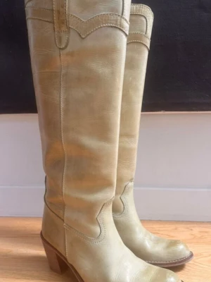 Beige höga cowboy boots i läder - Säljer ett par höga cowboy boots från Dico Copenhague i beige läder. Klassisk western-design med dekorativa sömmar och rundad tå. Bootsens skaft har dragflikar upptill och en stadig klack som ger extra attityd till din outfit. Passar fin med kort kjolar eller jeans Skinn 