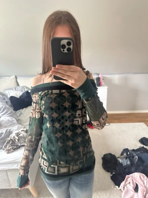 Grön mesh offshoulder topp - Säljer en grön mesh topp med offshoulder-design och lång ärm. Toppen har ett unikt geometriskt mönster i beige och svart, och är lätt transparent. Perfekt för dig som vill sticka ut med en cool och trendig stil.